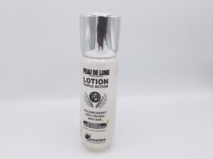 A bottle of PEAU DE LUNE LOTION TRIPLE ACTION ÉCLAIRCISSANT ANTI-TACHES ANTI-ÂGE AU GLUTA-C+ HUILES ESSENTIELLES 100 ml with text detailing its anti-spot and anti-aging properties, displayed against a white background.